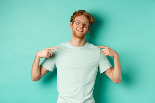 Fiducioso uomo dai capelli rossi con gli occhiali e la maglietta, sorridente con il viso compiaciuto e indicando se stesso, vantandosi in piedi su sfondo turchese.