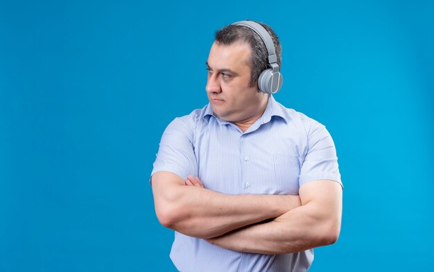 Fiducioso uomo di mezza età in camicia a righe blu che indossa le cuffie con le braccia piegate guardando al lato destro su uno spazio blu