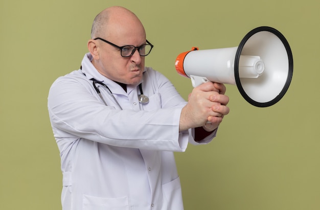 Fiducioso uomo slavo adulto con occhiali ottici in uniforme da medico con stetoscopio che tiene in mano un altoparlante che guarda di lato