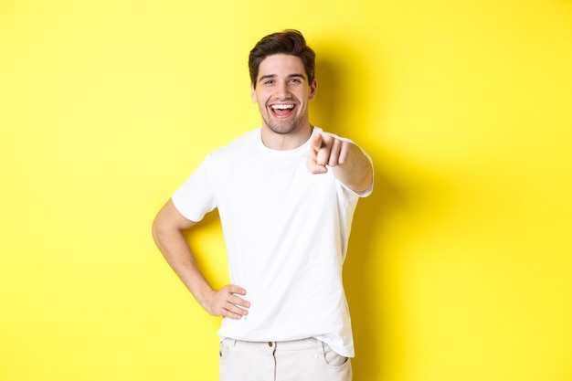 Fiducioso uomo sorridente che punta alla telecamera, in piedi in abiti bianchi su sfondo giallo.