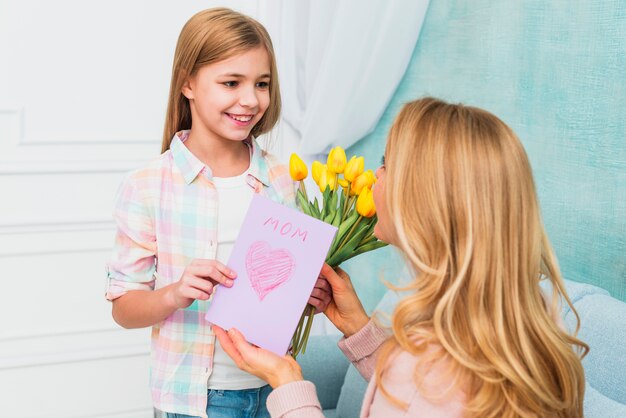Figlia che sorride e che presenta fiori e cartolina del giorno della madre per la mamma