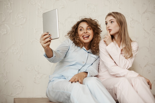 Figlia e madre che fanno foto di famiglia mentre si rilassano a casa. Bella mamma emotiva che prende selfie con cute bestie mentre entrambi in indumenti da notte, facendo facce e shh gesti come se avessero un segreto