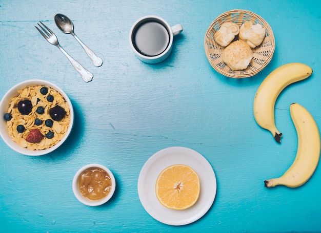 Fiocchi di mais; marmellata; arancia dimezzata; pane; caffè; banana sul contesto in legno blu