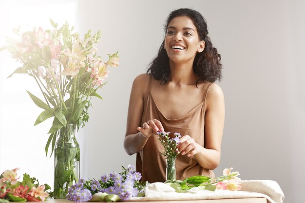 Fiorista di bella donna africana allegra sorridente che fa mazzo nel negozio di fiori sopra la parete bianca.