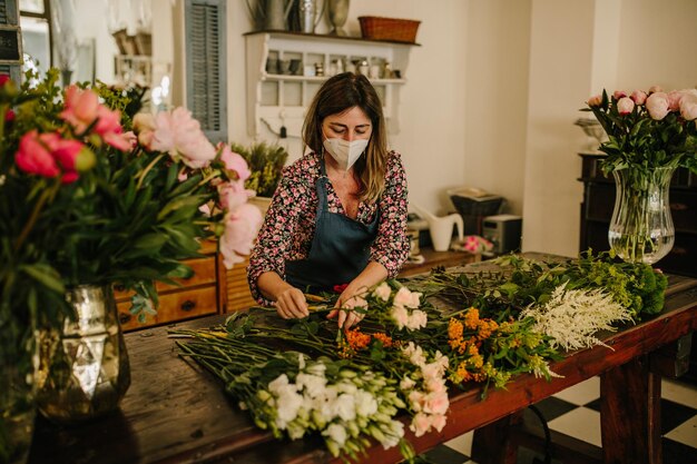 Fiorista europea con una maschera medica per il viso che fa composizioni floreali in uno studio di design floreale