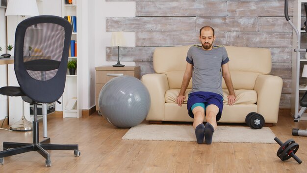 Fit guy training tricipiti sul divano di casa indossando abbigliamento sportivo.