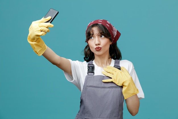 Flirty giovane donna addetta alle pulizie che indossa bandana uniforme e guanti di gomma che allungano il telefono cellulare tenendo la mano sul petto facendo gesto di bacio prendendo selfie isolato su sfondo blu