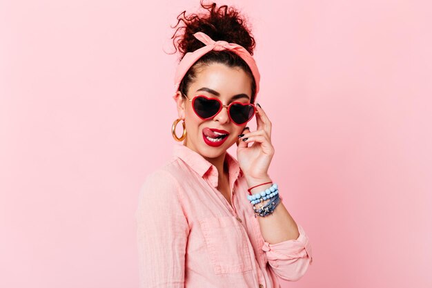 Flirty pinup girl con gli occhiali lecca le labbra rosse Istantanea di una donna con enormi orecchini e abito rosa in posa su sfondo isolato