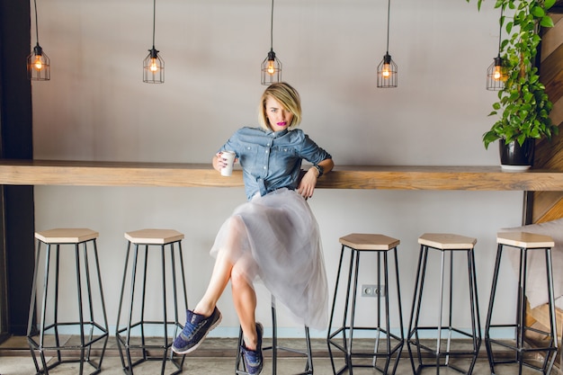 Flirty ragazza bionda con gli occhi azzurri e le labbra rosa brillante seduto in una caffetteria su una sedia a bere caffè.