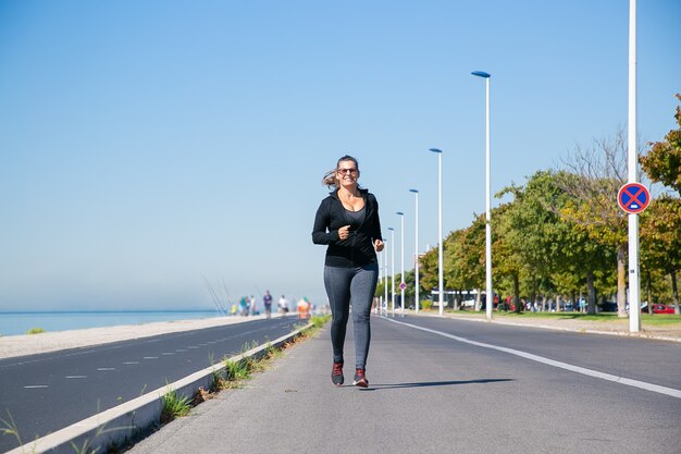 Focalizzato donna matura in abiti fitness fare jogging lungo la riva del fiume al di fuori, godendo la corsa mattutina. Vista frontale a figura intera. Concetto di stile di vita attivo