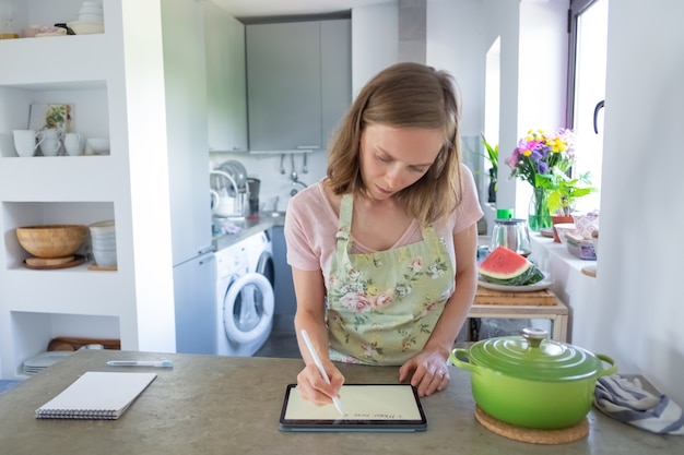 Focalizzato giovane donna pianificazione menu settimanale, scrivendo sullo schermo pad mentre cucina nella sua cucina, utilizzando tablet vicino alla grande pentola sul bancone. Vista frontale. Cucinare a casa e il concetto di famiglia