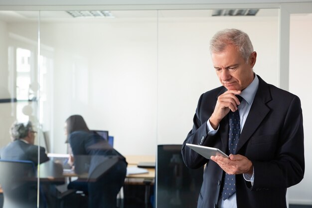 Focalizzato imprenditore pensieroso fissando lo schermo del tablet mentre i suoi colleghi discutono del progetto sul posto di lavoro dietro la parete di vetro. Copia spazio. Concetto di comunicazione