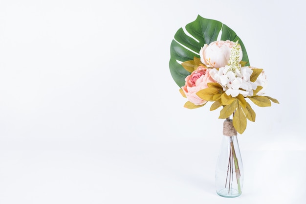 Foglia e fiori artificiali in vaso di vetro su bianco.