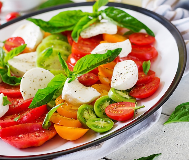 Foglie della mozzarella, dei pomodori e dell'erba del basilico in piatto sulla tavola di legno bianca. Insalata caprese. Cibo italiano.