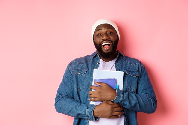 Formazione scolastica. Felice studente maschio afroamericano in berretto che tiene quaderni, studiando corsi, sorridendo alla telecamera, in piedi su sfondo rosa