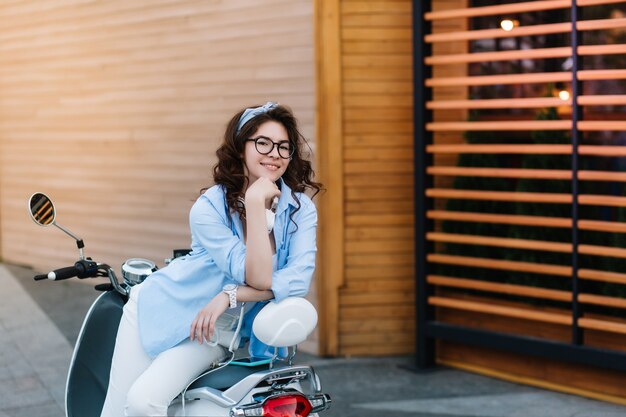 Formosa ragazza dai capelli scuri con un sorriso giocoso in posa sul ciclomotore e guardando con interesse