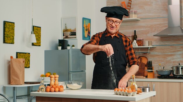 Fornaio esperto che sparge farina nella cucina di casa per la preparazione del cibo. Chef anziano in pensione con bonete e grembiule che cosparge, setacciando setacciando gli ingredienti crudi a mano che cuoce pizza fatta in casa, pane.