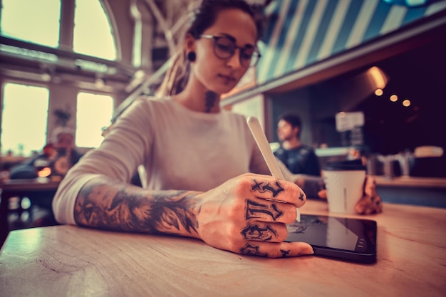 Foto a basso angolo di attraente ragazza pensosa con dreadlocks e tatuaggi. Dhe sta disegnando mentre è seduto al bar.