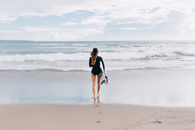 Foto a figura intera dal retro della giovane donna attraente con i capelli lunghi vestita in costume da bagno corre nell'oceano con una tavola da surf, sullo sfondo dell'oceano, sport, stile di vita attivo