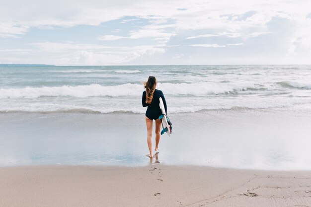 Foto a figura intera dal retro della giovane donna attraente con i capelli lunghi vestita in costume da bagno corre nell'oceano con una tavola da surf, sullo sfondo dell'oceano, sport, stile di vita attivo