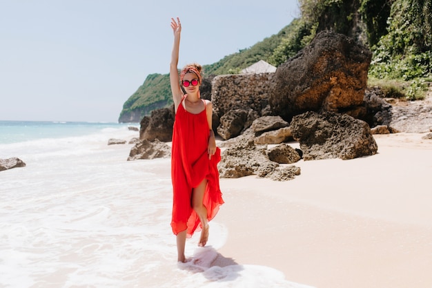 Foto a figura intera del ballo divertente della ragazza caucasica sveglia alla spiaggia del mare. La donna raffinata indossa un vestito rosso agghiacciante in spiaggia selvaggia al mattino.