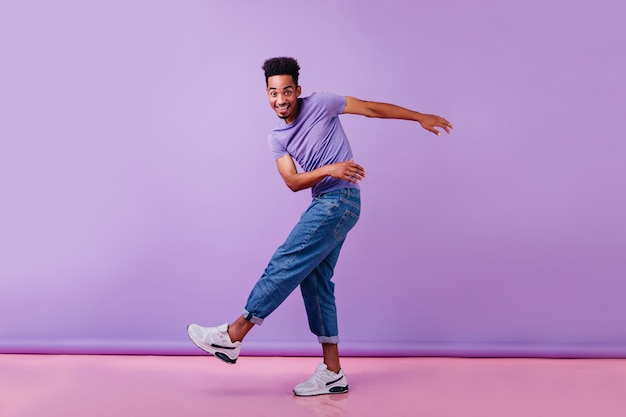 Foto a figura intera dell'uomo africano felice che balla con un sorriso sincero. Modello maschile in jeans e maglietta viola divertendosi.