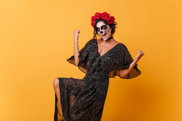 Foto a figura intera della ragazza sorridente che fa il gesto vincente. Signora in abito di chiffon nero posa con la maschera di Halloween.