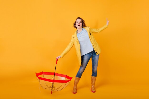 Foto a figura intera di donna sottile in abito autunnale che balla in scarpe di gomma. Attraente ragazza con capelli ondulati scherzare dopo la pioggia.