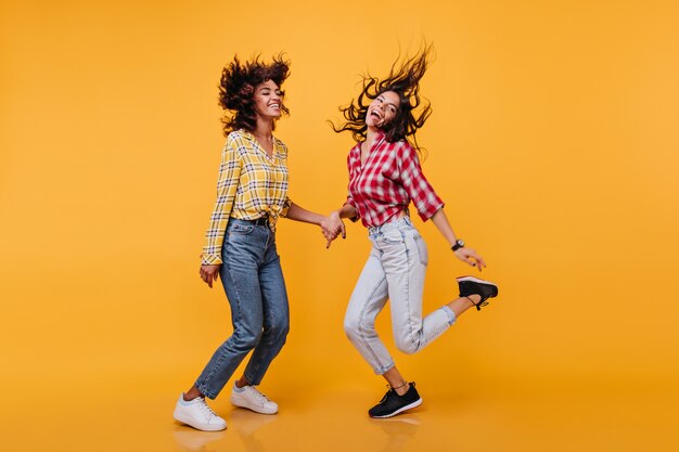 Foto a figura intera di giovani donne felici che si muovono con gioia. Le modelle dai capelli castani si tengono per mano e ridono in jeans di mamme