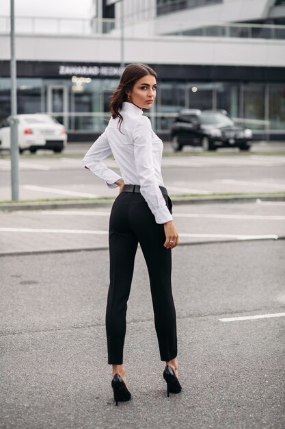 Foto a figura intera di una donna elegante vestita con pantaloni neri e camicia bianca e in piedi per strada contro l'edificio moderno. Stile e concetto di moda
