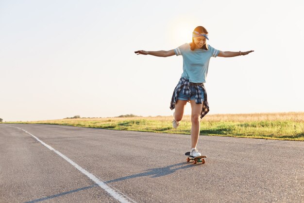 Foto a tutta lunghezza di una donna felice e attraente magra con emozioni positive e sorriso a trentadue denti, che cavalca skateboard in strada, trascorrendo l'estate in modo attivo.