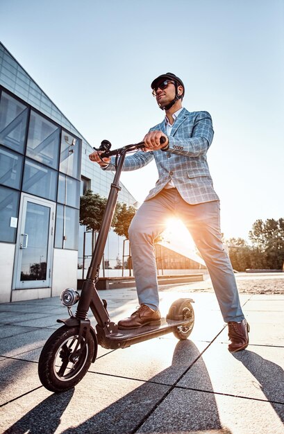 Foto ad angolo basso di un uomo elegante e intelligente con occhiali da sole e casco che guida il suo scooter elettrico.