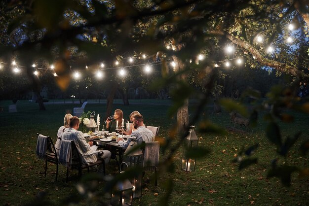 Foto attraverso i rami degli alberi con foglie. Tempo di sera. Gli amici cenano nello splendido posto all'aperto