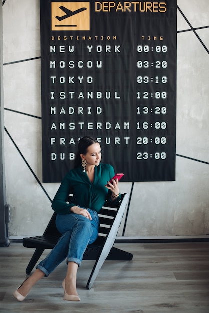 Foto d'archivio a figura intera di elegante donna d'affari in smart casual utilizzando il telefono cellulare in attesa del suo volo contro il poster della partenza.