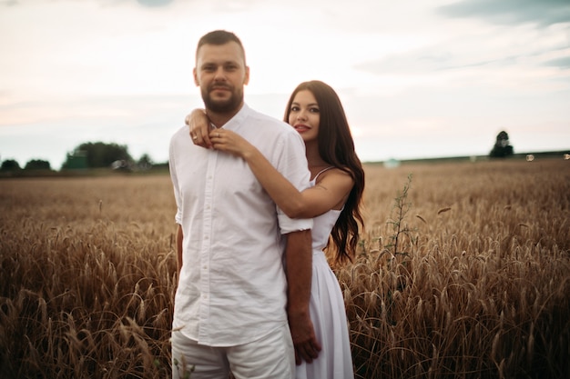 Foto d'archivio ritratto di un ragazzo barbuto che abbraccia la sua splendida ragazza entrambi in abiti bianchi che si abbracciano nel campo di grano. Bellissimo campo di grano in background.