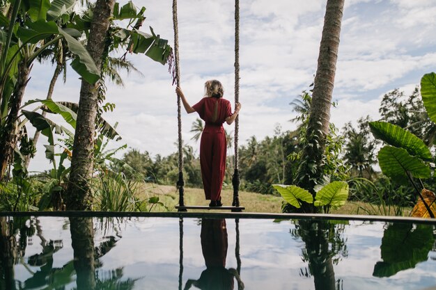 Foto dal retro della donna sottile in abito lungo guardando il cielo piovoso. Colpo esterno del modello femminile formoso che gode della vista della natura al resort.