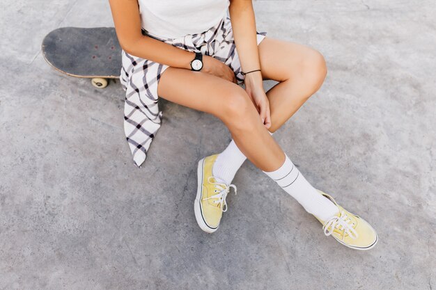 Foto dall'alto della donna caucasica abbronzata agghiacciante in skate park dopo l'allenamento. Magnifica donna indossa calzini bianchi e scarpe gialle seduto sullo skateboard.