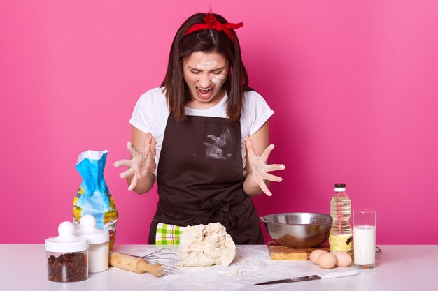 Foto del panettiere castana arrabbiato che è malato e stanco della pasta d'impasto, vestito in maglietta casuale e grembiule marrone sporchi con farina. La femmina allarga le dita mentre grida qualcosa. Concetto di cibo.