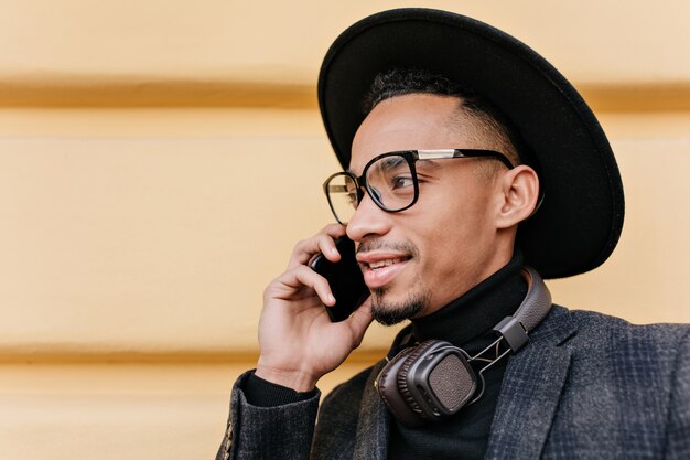 Foto del primo piano del modello maschio americano in cappello nero. Ritratto all'aperto di bell'uomo africano che parla al telefono sulla strada al mattino.