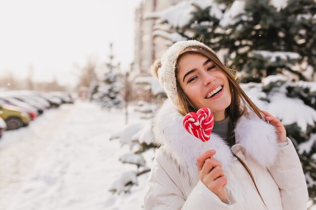 Foto del primo piano dell'incantevole donna dai capelli lunghi che cammina sulla strada innevata con lecca-lecca. Donna graziosa che ride in cappello lavorato a maglia che gode del fine settimana invernale all'aperto.