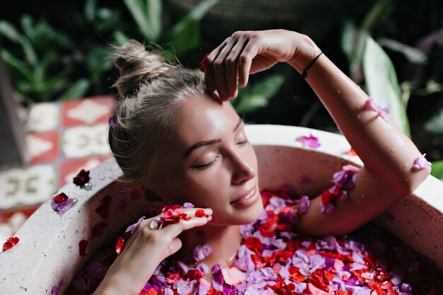 Foto del primo piano della donna abbronzata attraente agghiacciante nella vasca piena di petali di rosa. Colpo sopraelevato dell'interno del modello femminile biondo raffinato che gode della stazione termale con un sorriso dolce.