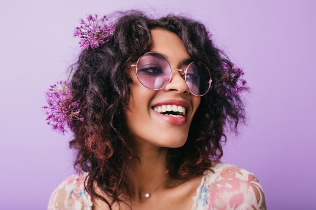 Foto del primo piano della signora africana di buon umore con fiori nei capelli. Tiro al coperto di ridere ragazza nera isolata.