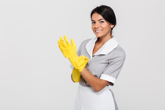 Foto del primo piano di giovane domestica castana sorridente in uniforme che indossa i guanti di gomma gialli mentre stando