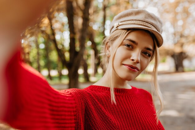Foto del primo piano di un'affascinante bionda in maglione rosso che fa bella selfie nella sosta di autunno. Splendida giovane donna con trucco naturale in posa all'aperto in elegante cappello leggero.