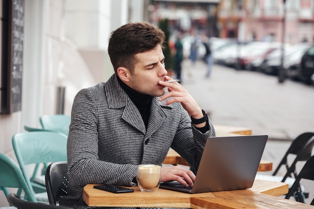 Foto dell'uomo bello in sigaretta di fumo del cappotto grigio e bere cappuccino mentre riposando in caffè all'aperto