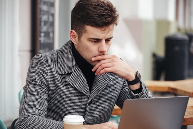 Foto dell'uomo d'affari che lavora con il computer portatile d'argento in caffè fuori, bevendo caffè da asporto