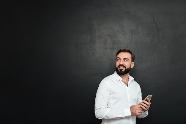 Foto dell'uomo felice in camicia bianca che guarda indietro mentre chiacchierando o usando internetin senza fili sul telefono cellulare sopra grigio scuro