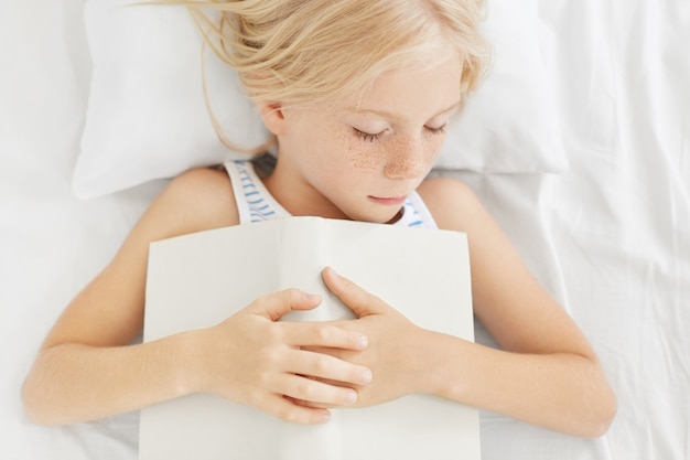 Foto della bambina bionda con le lentiggini che sonnecchia nel letto, tenendo il libro in mano, sentendo stanchezza dopo una lunga lettura, addormentarsi. Ragazza sonnolenta calma che si trova sui vestiti bianchi del letto con il libro.