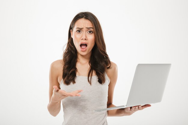 Foto della donna del brunette che osserva sulla macchina fotografica perplesso e che tiene taccuino d'argento a disposizione, isolato sopra la parete bianca