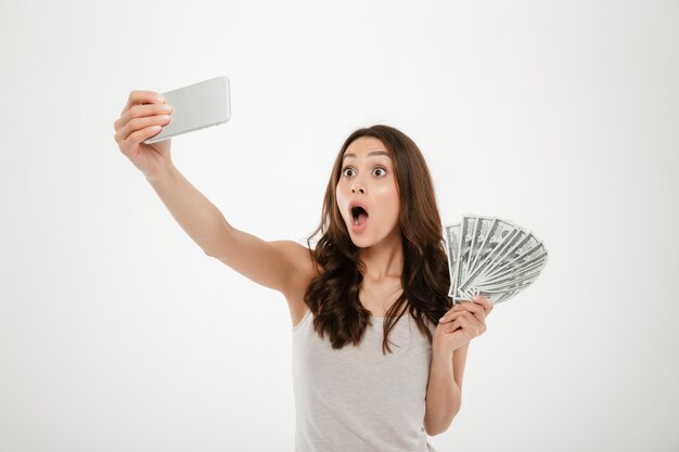 Foto della donna divertente colpita che fa selfie che fotografa sul cellulare d'argento, telefono mentre tenendo fan delle banconote in dollari isolate sopra la parete bianca
